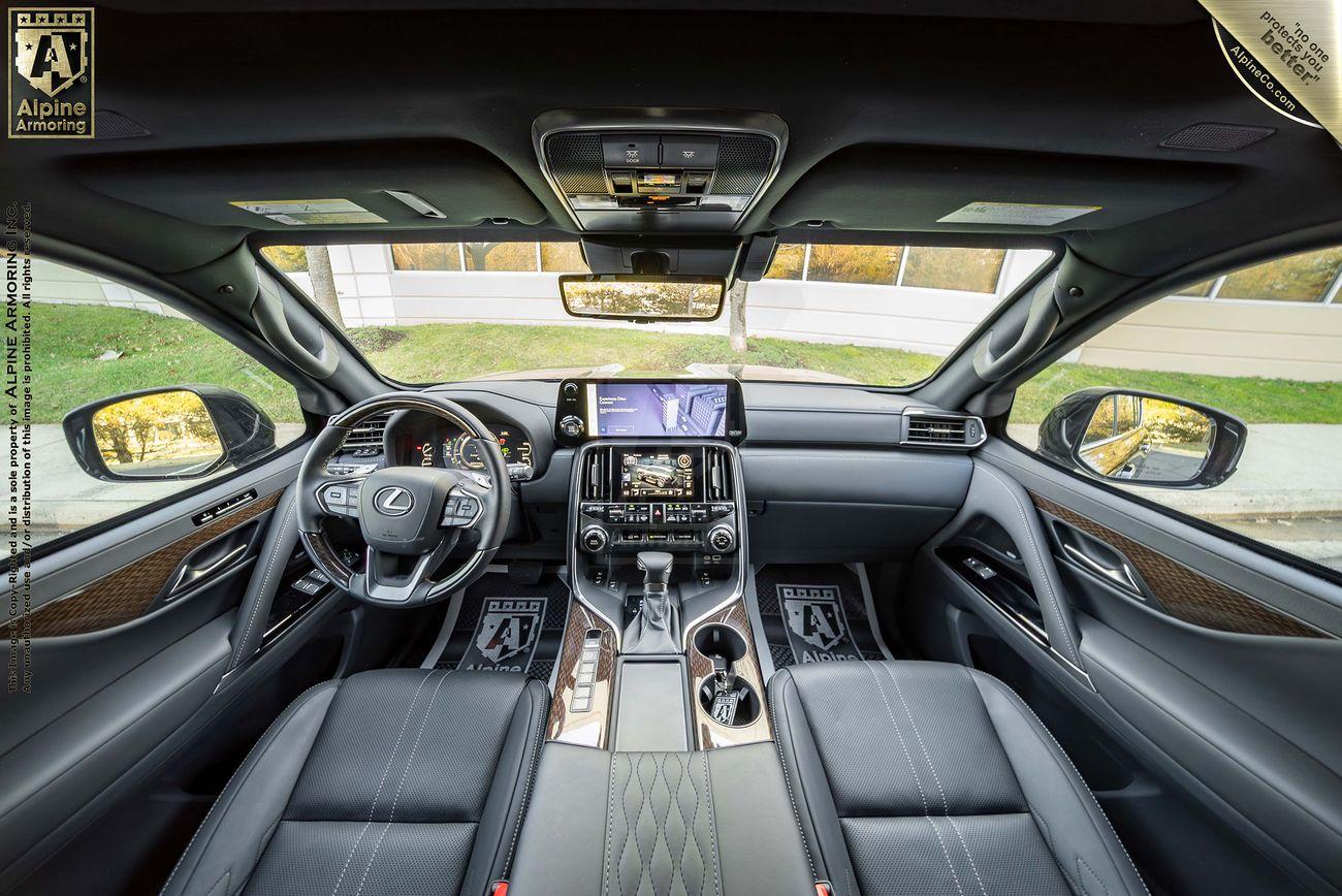 A view of the front interior of a armored Lexus LX600 Ultra Luxury SUV showing the dashboard, steering wheel, infotainment system, and leather seats. The Alpine Armoring logo is visible on the top left corner.