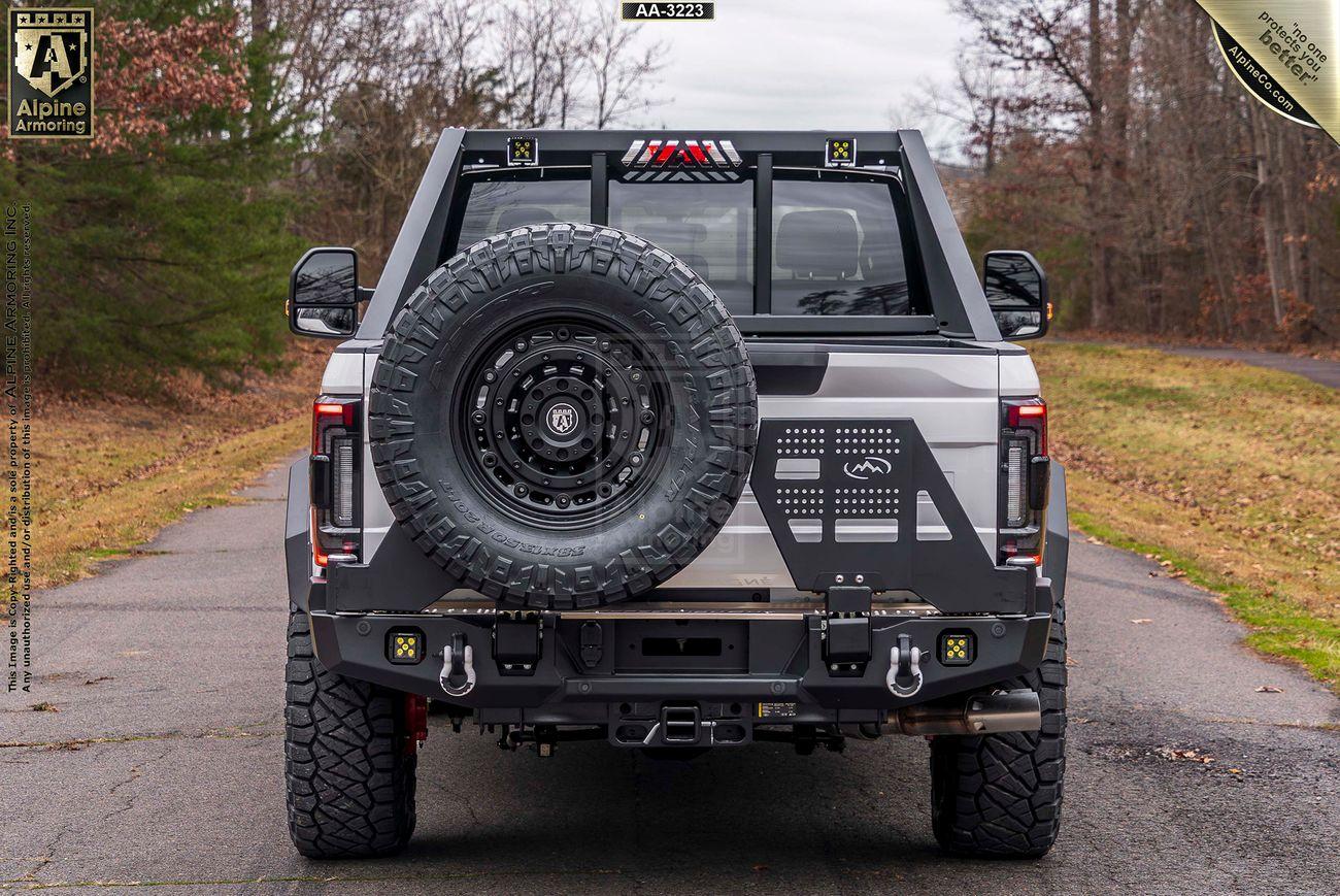 A Mastiff is shown from the rear view, featuring a spare tire mounted on the back and various equipment attached. The surroundings include a paved path with grassy and wooded areas.