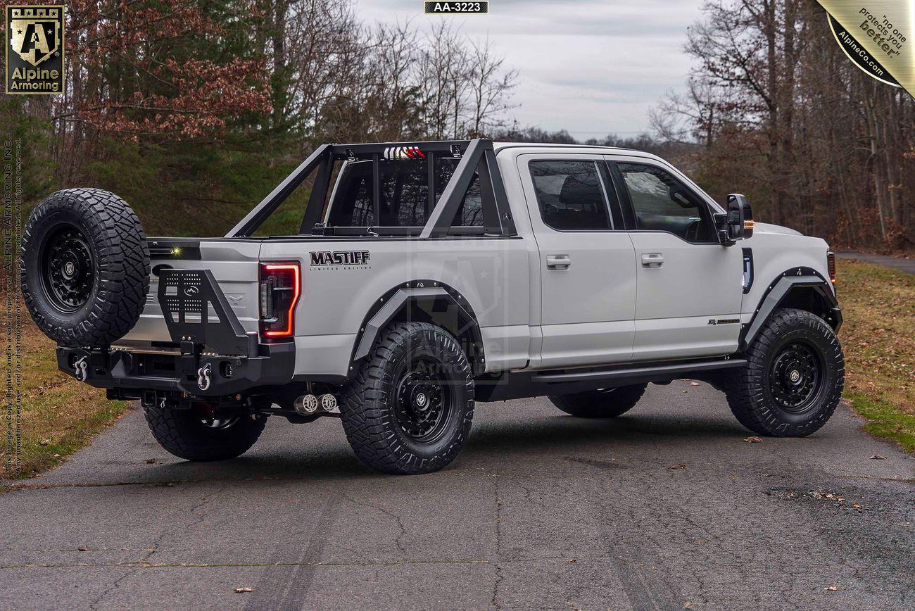 A white, heavily modified Mastiff truck with large tires and spare tire mounted in the rear stands on a paved road near wooded areas. The truck is branded with "Mastiff" and Alpine Armoring logos.