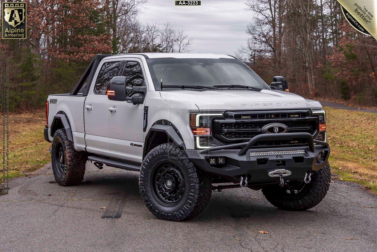 White Mastiff truck with off-road modifications, including large tires, a front bumper with a winch, and a roof rack, parked on a paved surface with trees in the background.