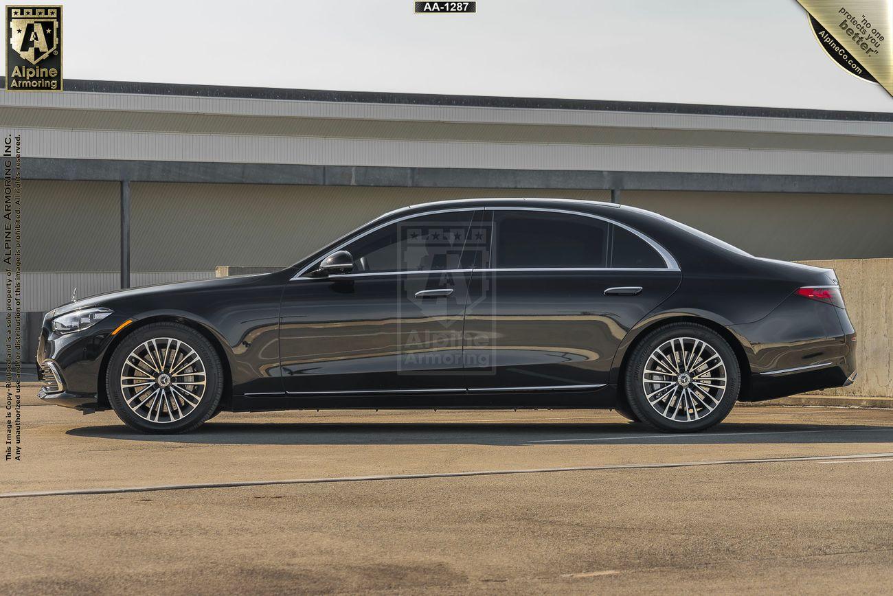 Driver-side profile of a black armored Mercedes-Benz S580 Executive sedan parked in an outdoor setting. The car features stylish rims and a sleek design. There is a warehouse in the background.