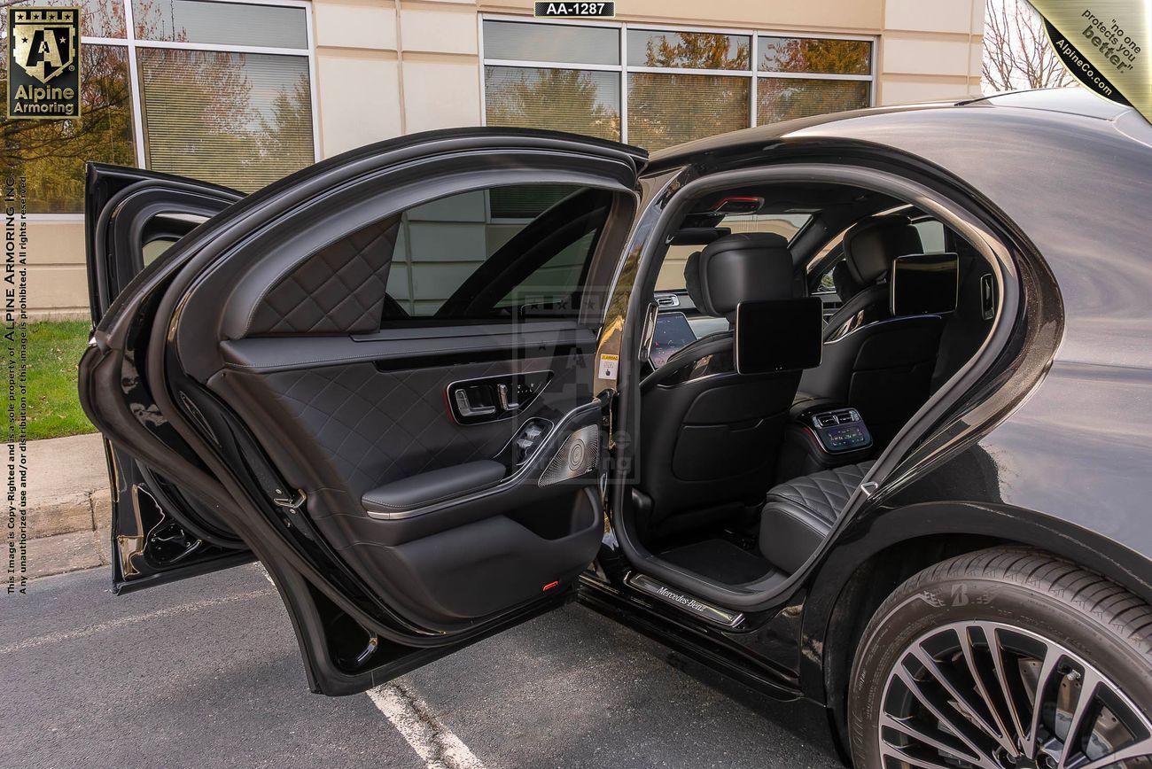 A black armored Mercedes-Benz S580 Executive car with open rear passenger doors showcasing a spacious, black leather interior. The vehicle is parked in an outdoor setting near a building with large windows.