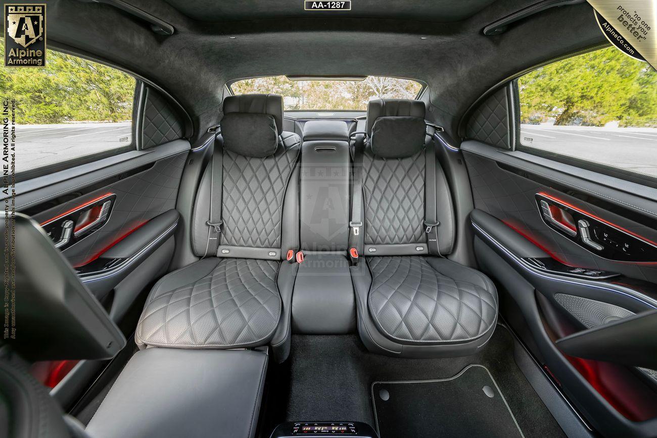 Interior of an armored Mercedes-Benz S580 Executive sedan featuring quilted leather rear seats, a central armrest with controls, and black and red accents. The rear window shows a tree-filled landscape outside.