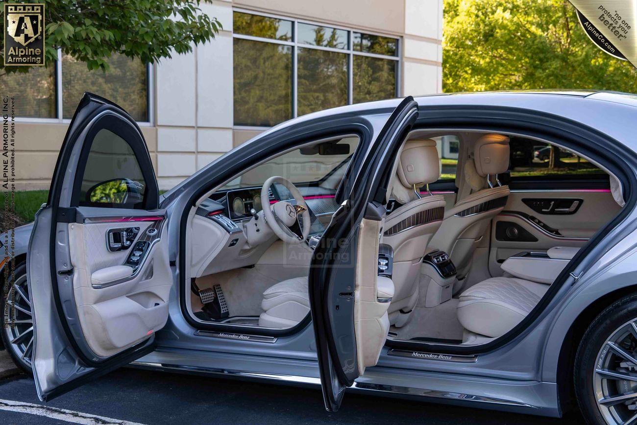 An armored silver Mercedes-Benz S580 sedan sedan with both front and back passenger doors open, revealing a cream-colored leather interior parked in a lot next to a building.