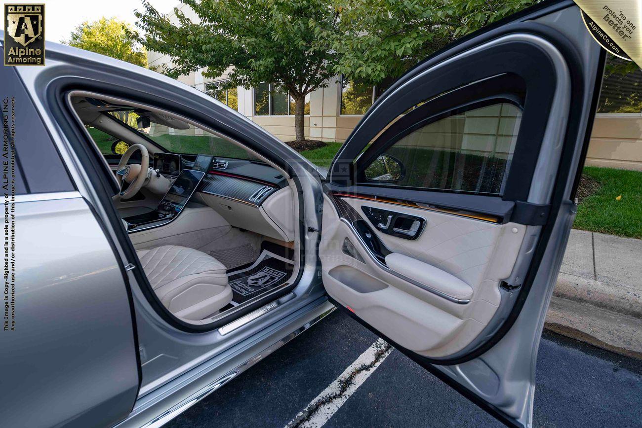 Open passenger side door of an armored silver Mercedes-Benz S580 sedan, displaying a spacious and elegant interior with light-colored upholstery and modern design features. Trees and a building are visible in the background.