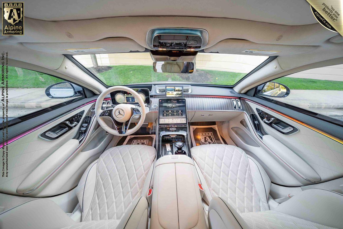 Interior of a Mercedes-Benz S580 sedan featuring white quilted leather seats, a central console with numerous controls, a wide dashboard with a touchscreen, and a sunroof. The steering wheel has a Mercedes logo.