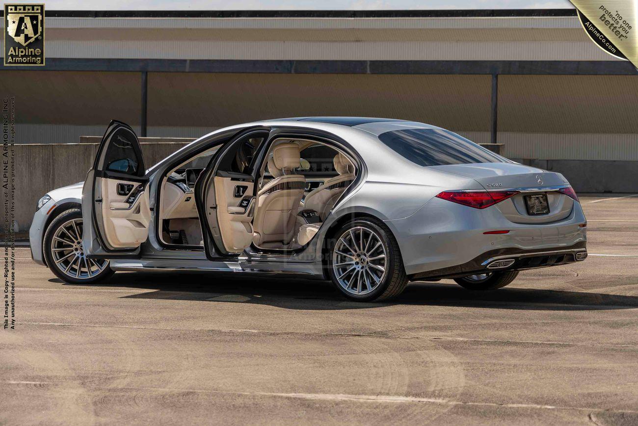 A silver Mercedes S580 sedan luxury sedan with open doors reveals beige interior seating. The car is parked in a mostly empty lot with a building in the background.