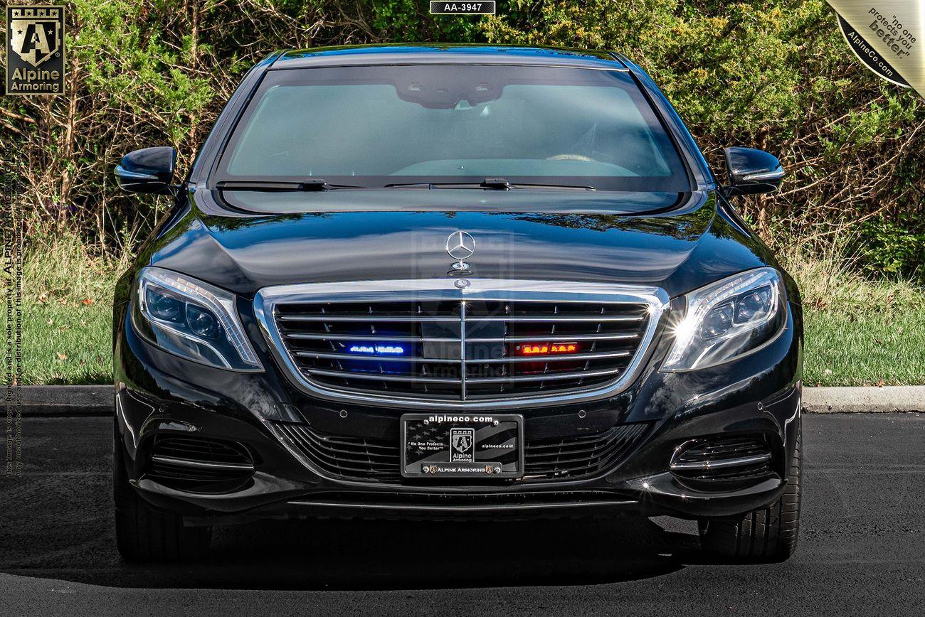 Front view of an armored black Mercedes-Benz S550 sedan  with police lights activated, displaying blue and red lights on the front grille. The car is parked on a road with greenery in the background.