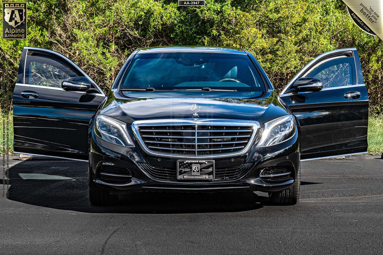 A black armored black Mercedes S550 luxury sedan with both front doors open, parked on a paved surface with green foliage in the background.