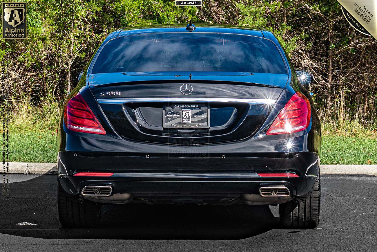 Rear view of a black Mercedes-Benz S-Class parked on a road with trees in the background. The car has a license plate frame with the "Alpine Armoring" logo.