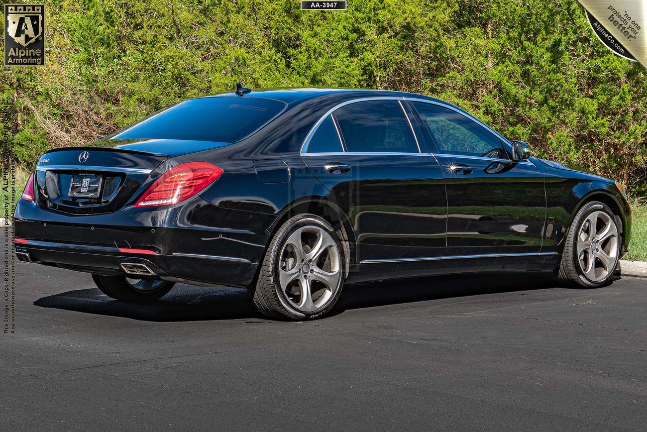 A black luxury armored black Mercedes S550 sedan is parked on a pavement with a background of green foliage.