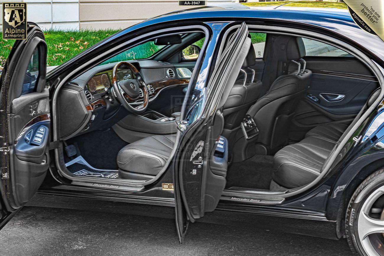 An armored black Mercedes S550 with all four doors open, revealing a sleek interior with leather seats and a wood-trimmed dashboard. The car is parked on a paved surface in an outdoor setting.