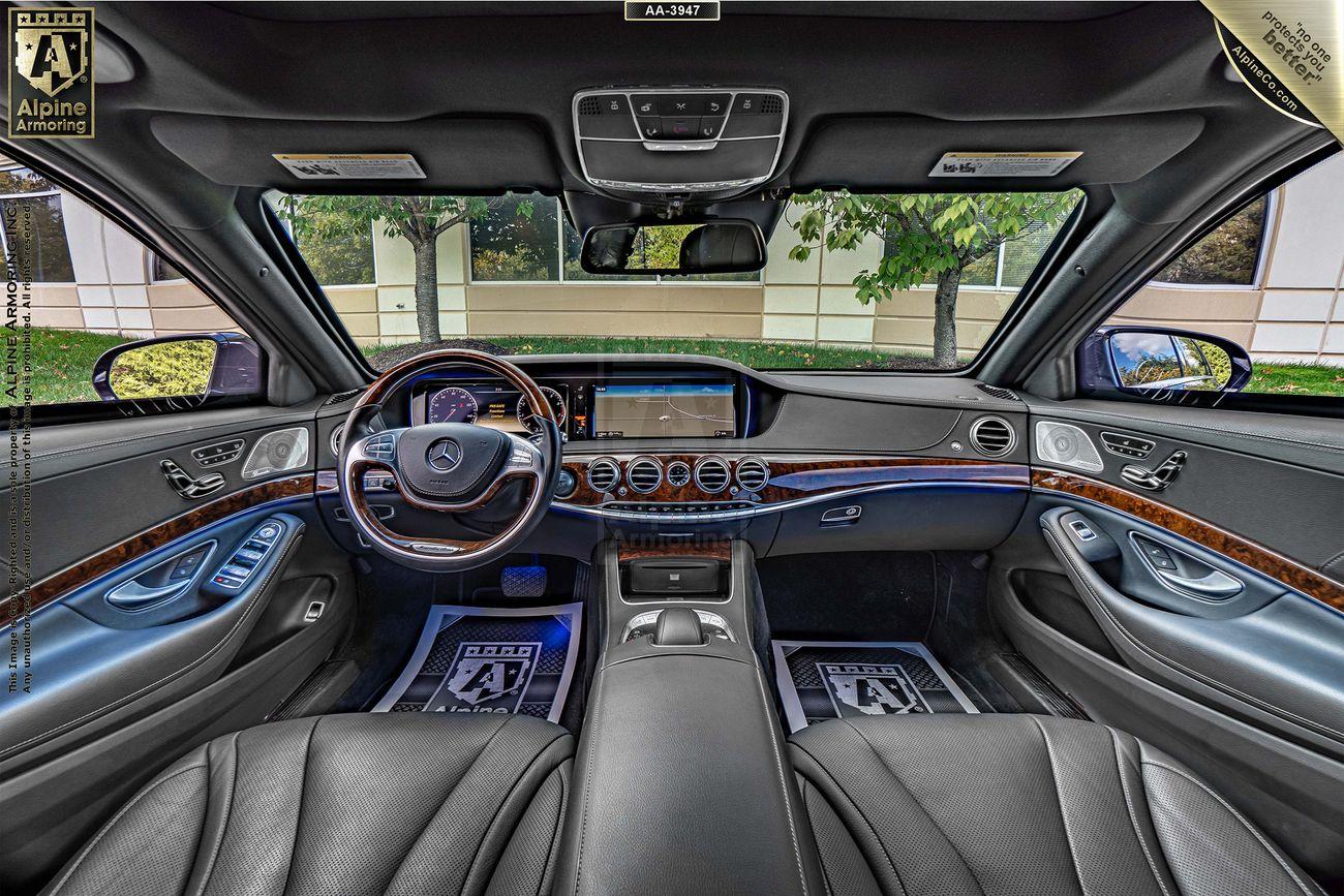 Interior view of armored black Mercedes S550 luxury sedan  featuring a leather-wrapped steering wheel, digital dashboard, wood trim accents, and two front seats separated by a central console. Trees visible through the windshield.