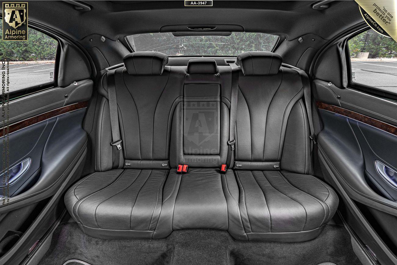 A view of an armored Mercedes S550's back seat, featuring black leather upholstery, three seats, two headrests, and a central armrest slot. The interior has wooden trim accents on the door panels.