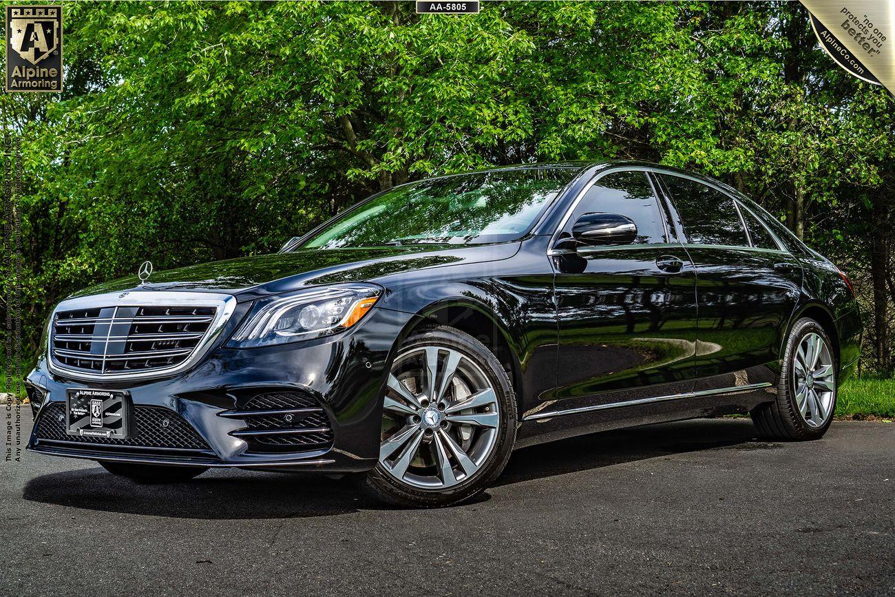 A black Mercedes-Benz S560 sedan is parked on a paved driveway with green foliage in the background. The car's emblem and grille are prominently visible.