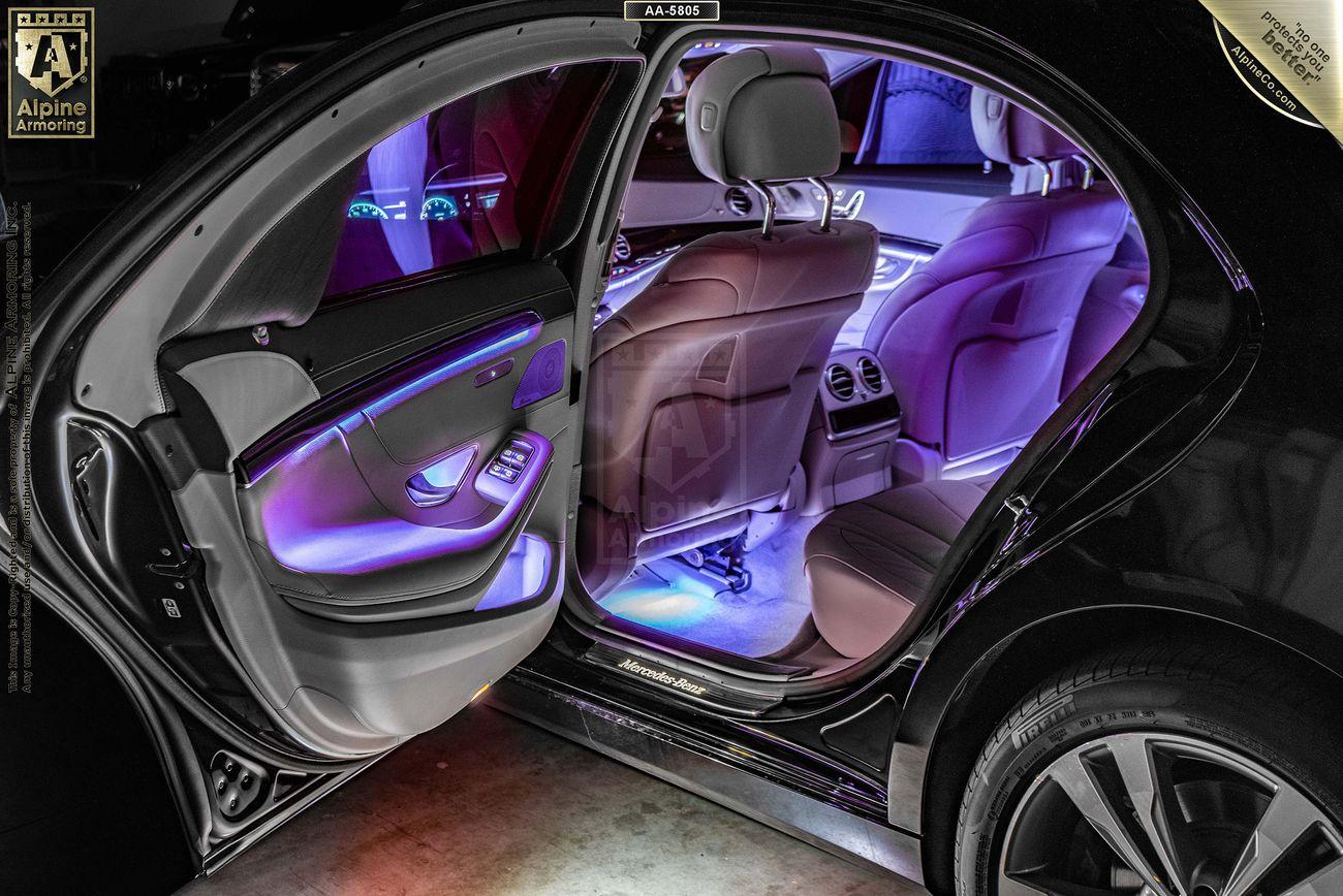 Open car door showing illuminated interior with purple and blue lights, visible dashboard with electronic displays, and front seats in a luxury vehicle.