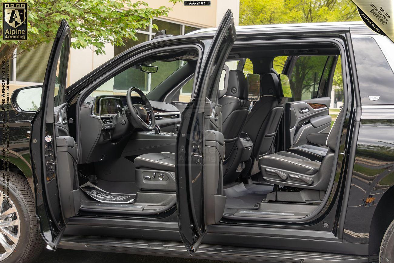 A black armored Cadillac Escalade ESV Premium Luxury SUV with all doors open, showcasing three rows of gray leather seats and a modern dashboard, parked outdoors with some greenery visible in the background.