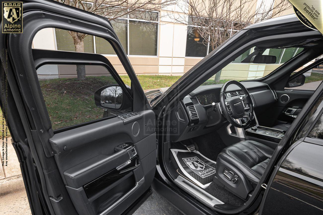 The interior of a black armored Range Rover Autobiography with the driver's side door open, showing black leather seats, steering wheel, and dashboard. A building and trees are visible in the background.