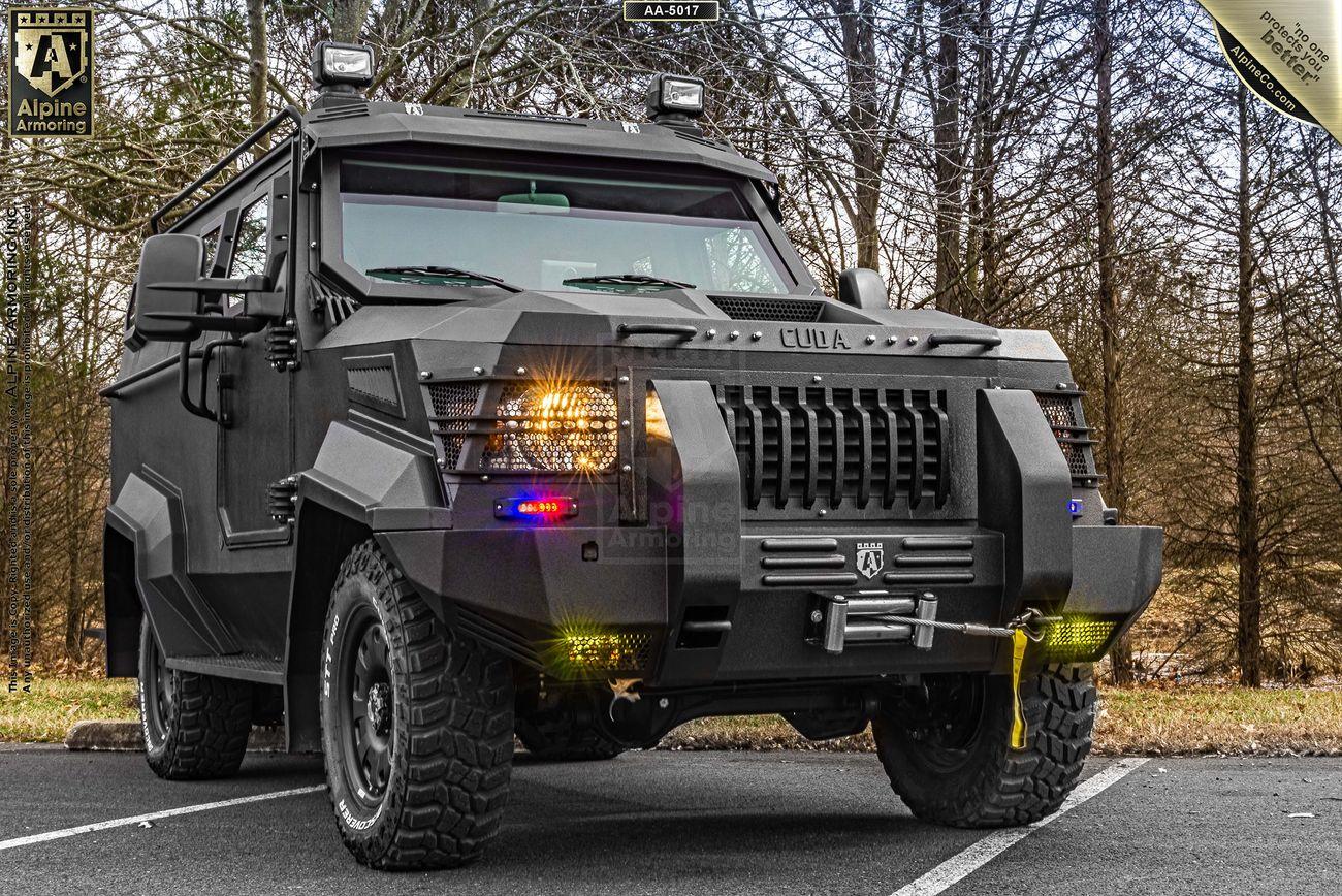 A black armored SWAT APC CUDA with rugged tires and a robust exterior is parked on a lot with trees in the background.