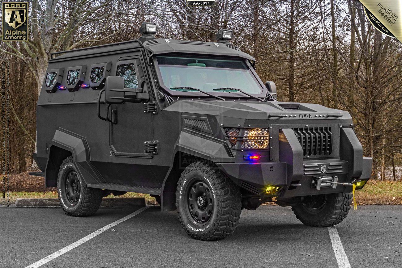 A black armored SWAT APC CUDAis parked on a paved surface in an outdoor setting with trees in the background. The vehicle features heavy-duty tires and multiple lights.