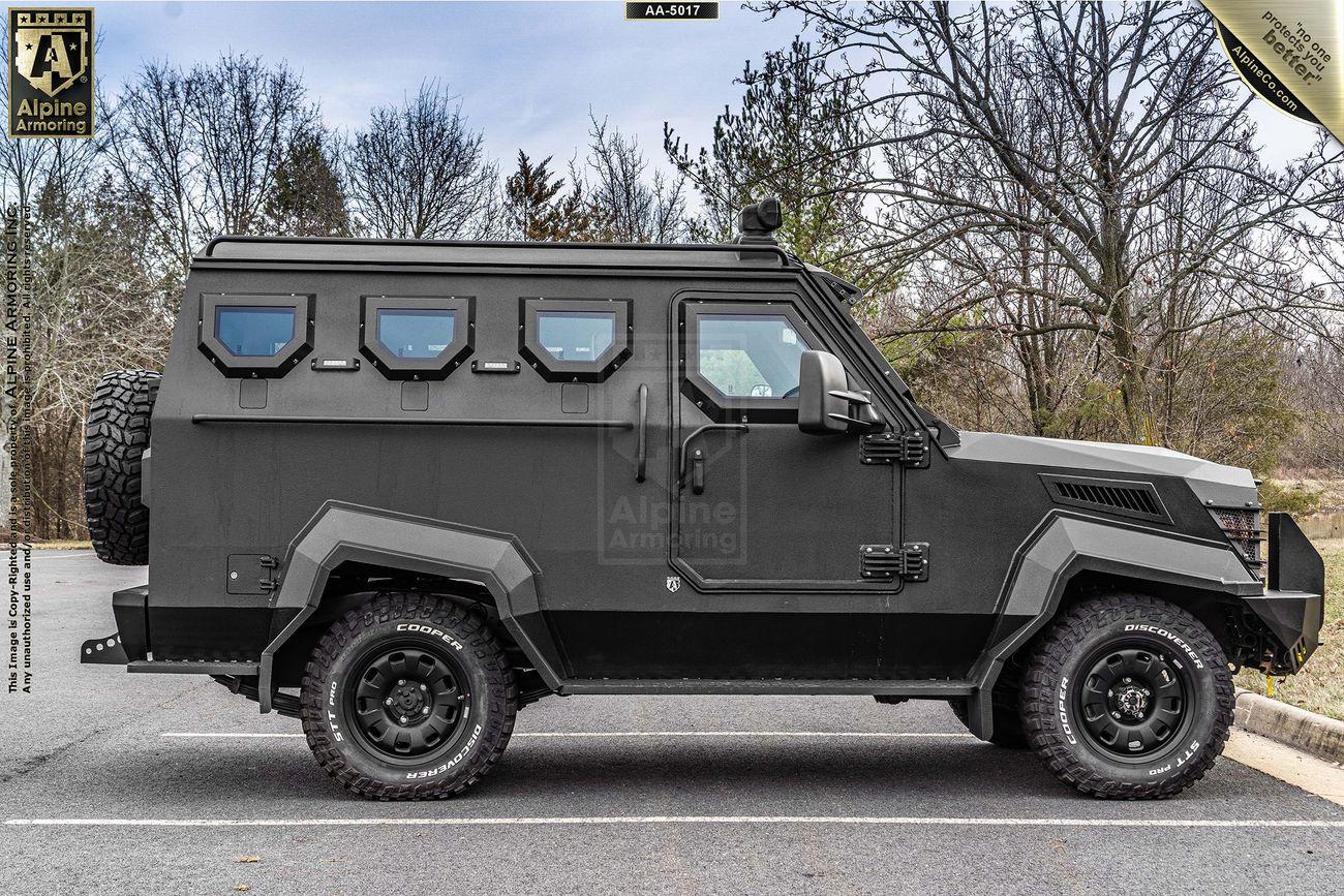 A matte black armored SWAT APC CUDA is parked on the street. It has six windows, rugged tires, and thick armored sides. Trees and a cloudy sky are in the background.