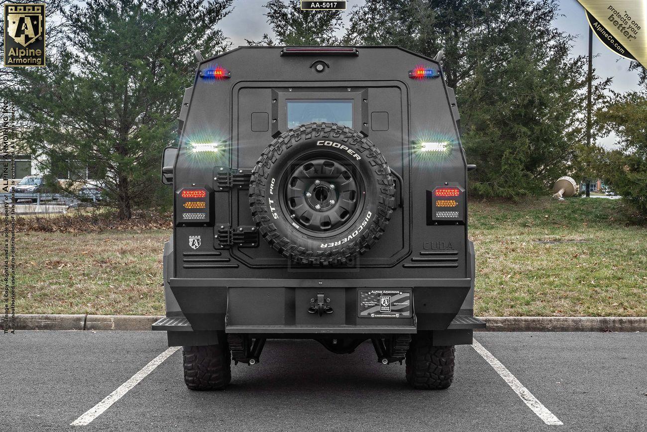 A rear view of an armored SWAT APC CUDA with a spare tire mounted on the back. The vehicle is parked in an outdoor area with trees in the background.