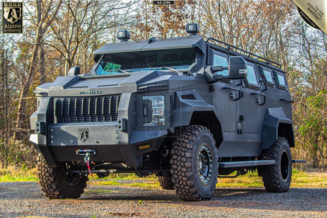 A tactical black armored Pit-Bull VX by Alpine Armoring stands on a gravel path surrounded by trees, featuring rugged tires, reinforced body, and advanced surveillance equipment on the roof.