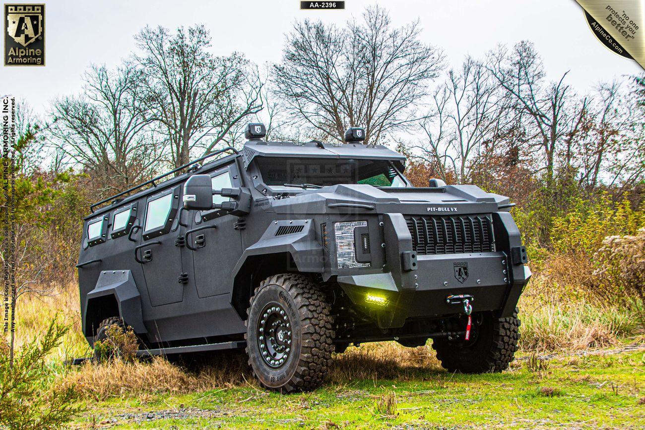 An armored Pit-Bull VX is parked on grass with trees in the background. The vehicle has a rugged design with large tires and is equipped with lights and winch.