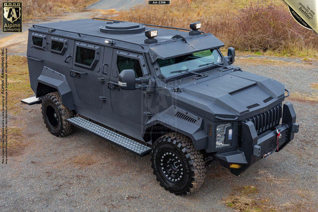 A black, heavily armored Pit-Bull VX is depicted parked on a gravel area with rugged terrain in the background. The vehicle has reinforced windows, heavy-duty tires, and appears designed for tactical use.