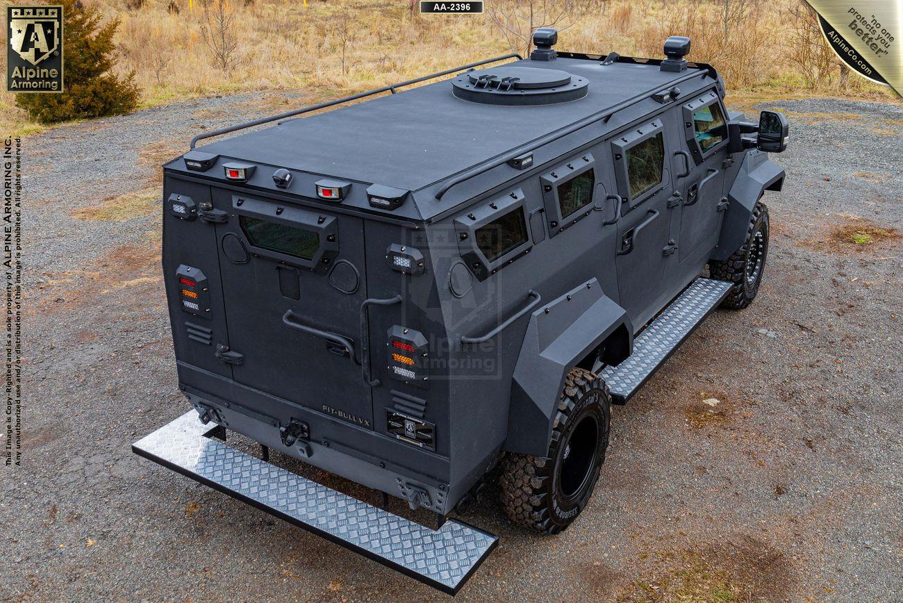 A matte black Pit-Bull VX is parked on a dirt road. The vehicle features heavy-duty tires, a reinforced body, and roof hatches, suggesting it is designed for rugged terrain and high-security purposes.