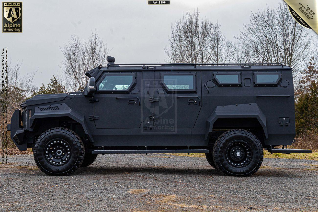 Side view of a black Alpine Armoring Inc. armored Pit-Bull VX parked on a gravel surface with leafless trees in the background.