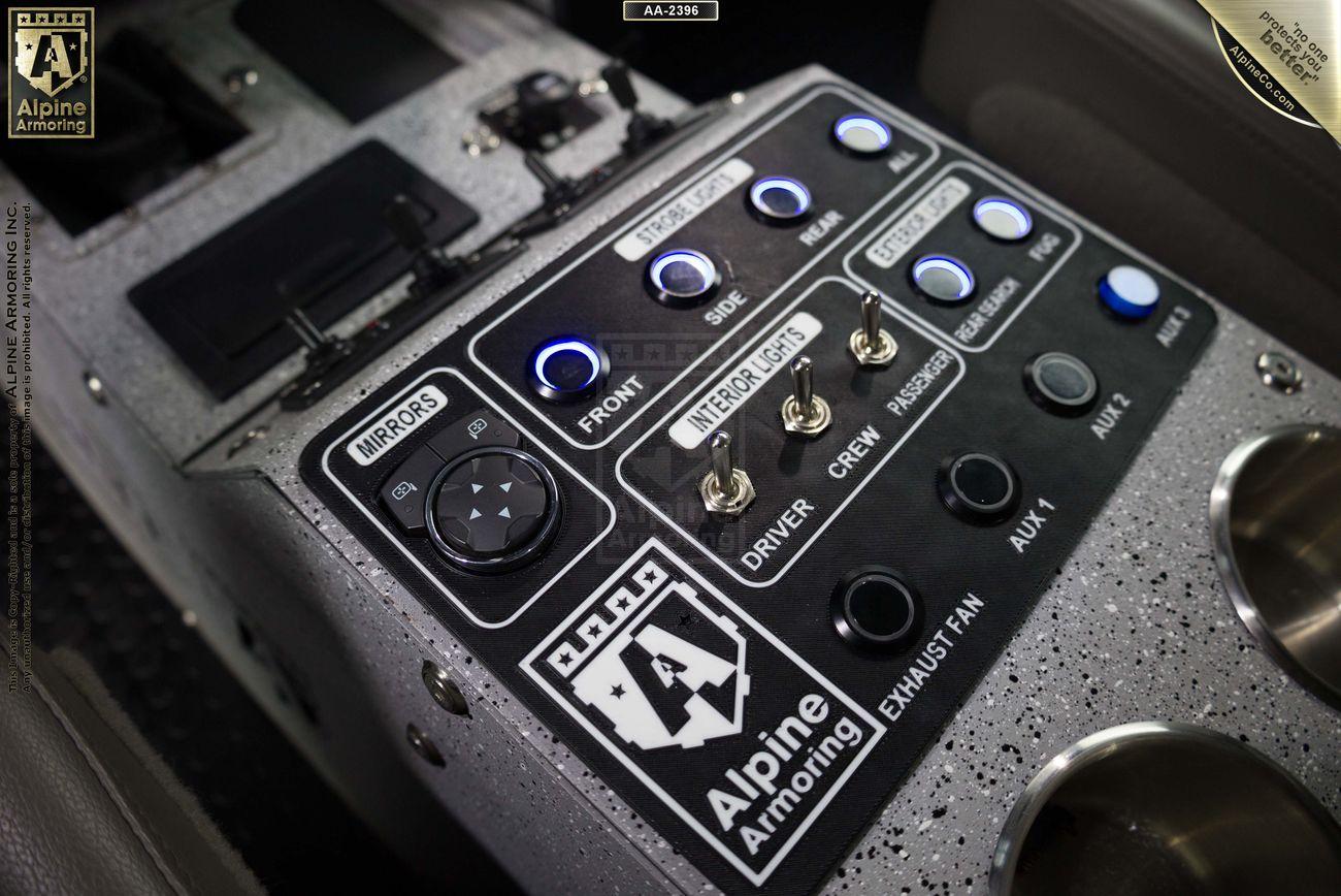 Close-up of a Pit-Bull VX control panel labeled "Alpine Armoring," featuring switches and knobs for functions like mirrors, intercom, lights, and exhaust fan, with cup holders on the right.