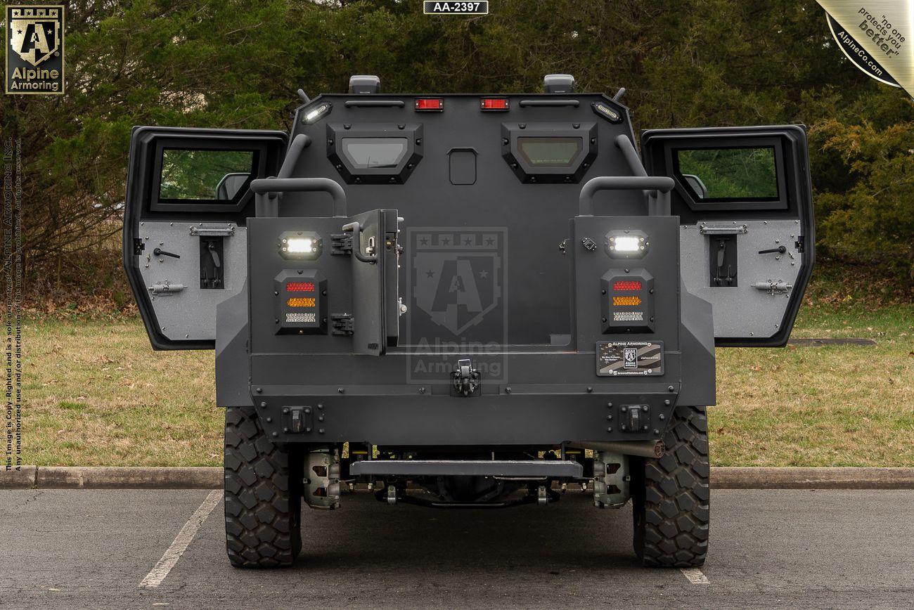 A gray, heavily armored black Pit-Bull VXT with two rear doors open, situated in a parking lot with trees in the background.