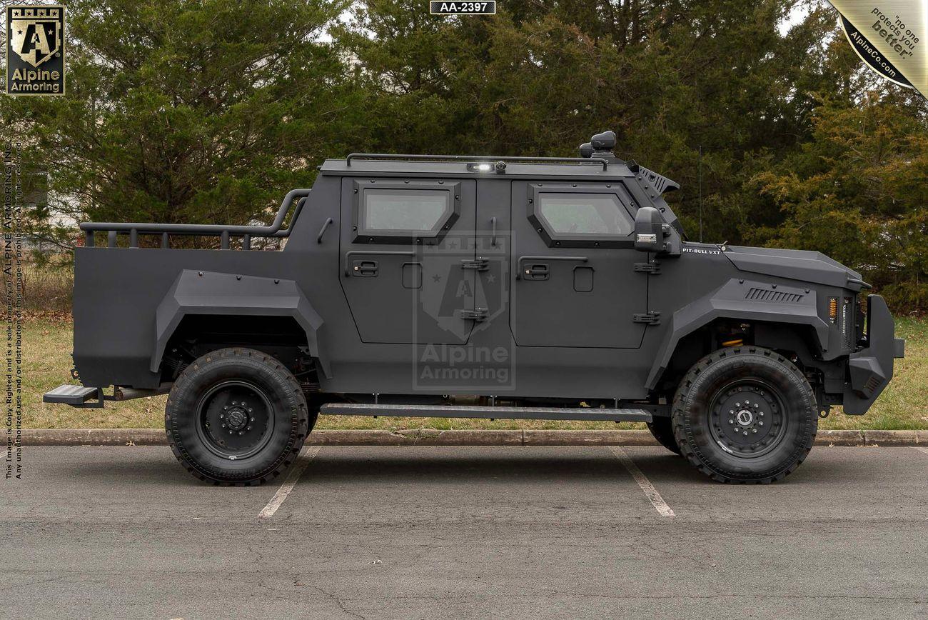 Side view of a black armored Pit-Bull VXT by Alpine Armoring, parked on a road with trees in the background.