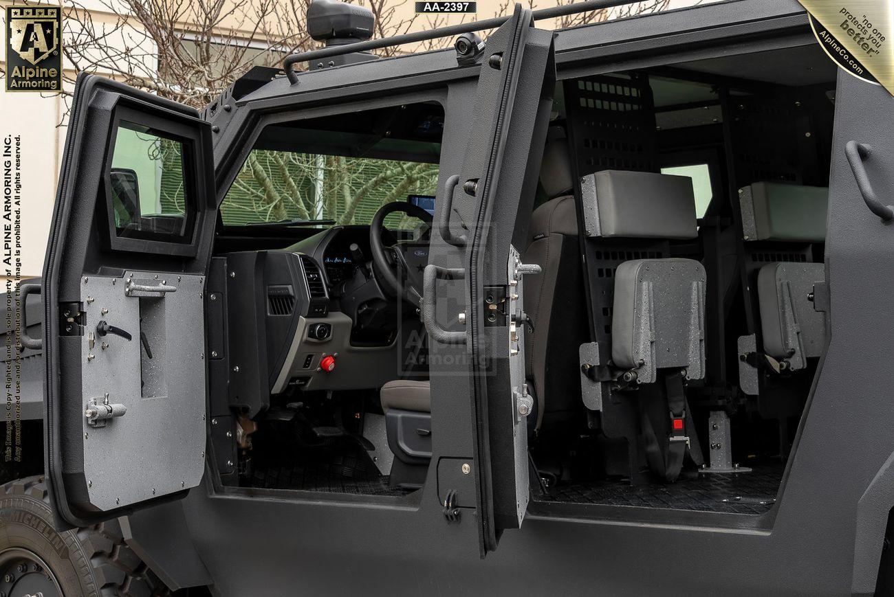 Interior view of an armored Pit-Bull VXT with the driver's door and rear side door open, revealing the driver's seat and passenger compartments. Black and gray colors dominate the vehicle's interior.