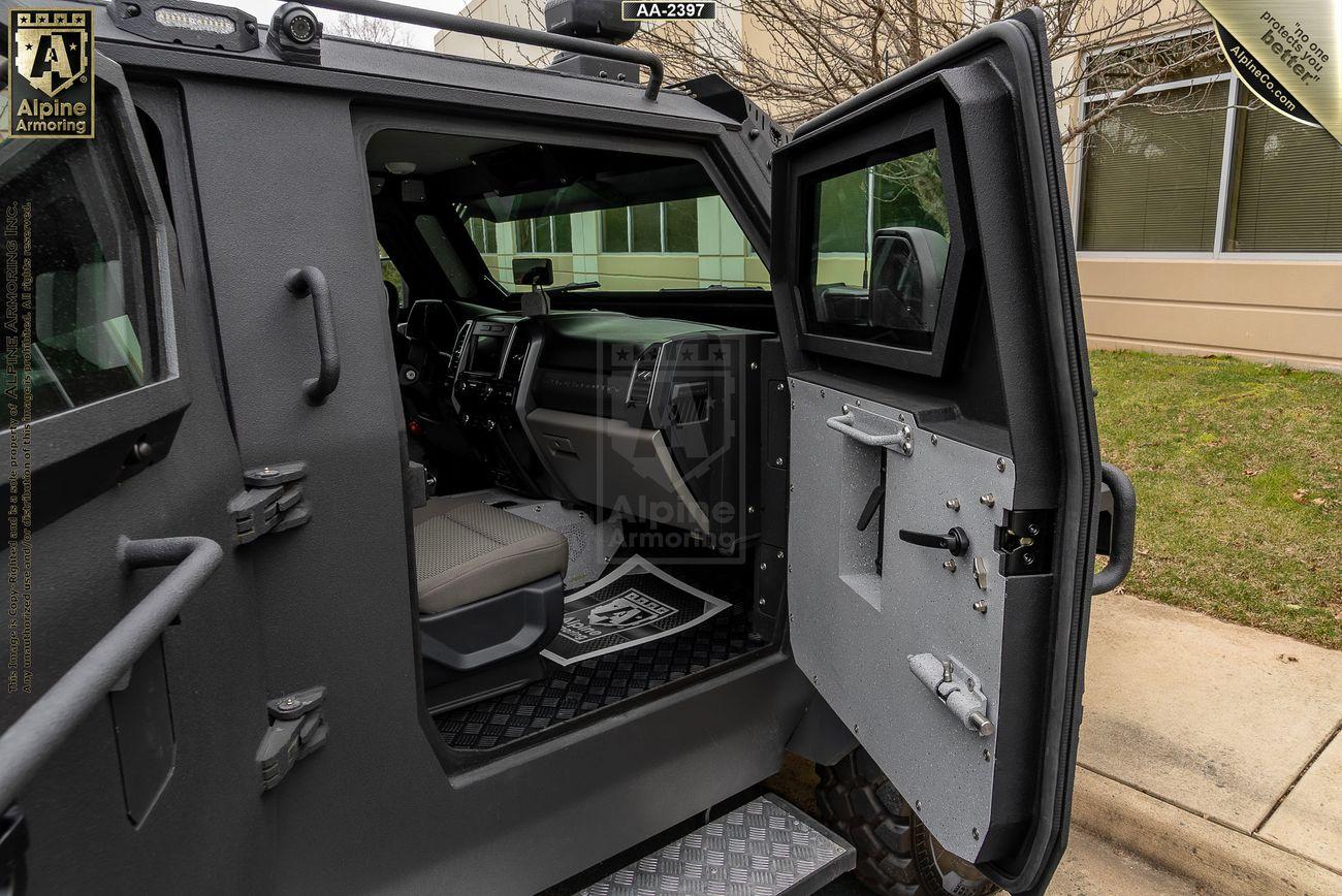 An armored black Pit-Bull VXT with an open driver's side door, showcasing the interior seating and dashboard. The exterior appears matte black, and a building is visible in the background.