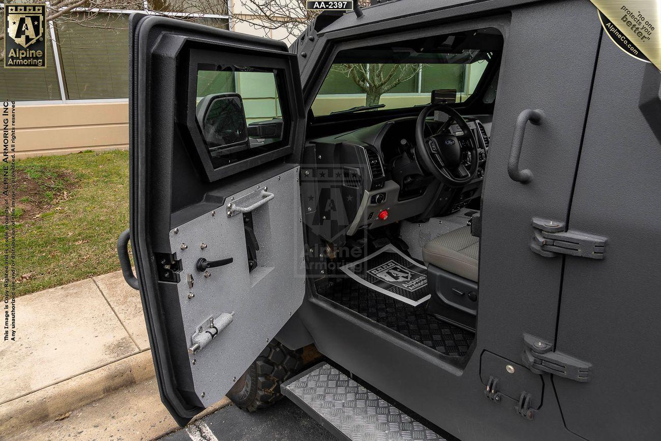 An open armored vehicle door from a black Pit-Bull VXT revealing the intricate interior with a steering wheel and digital dashboard. The surroundings suggest an outdoor setting with trees and buildings in view.