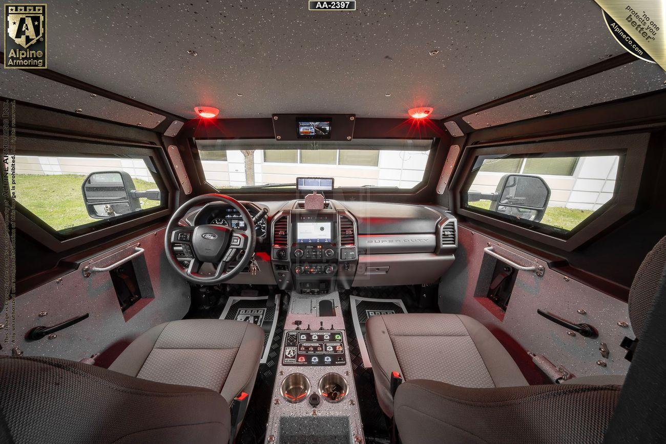 Interior view of a armored black Pit-Bull VXT's driver and passenger seats, dashboard, multiple control panels, and displays. The cabin is equipped with advanced instrumentation and red interior lights.