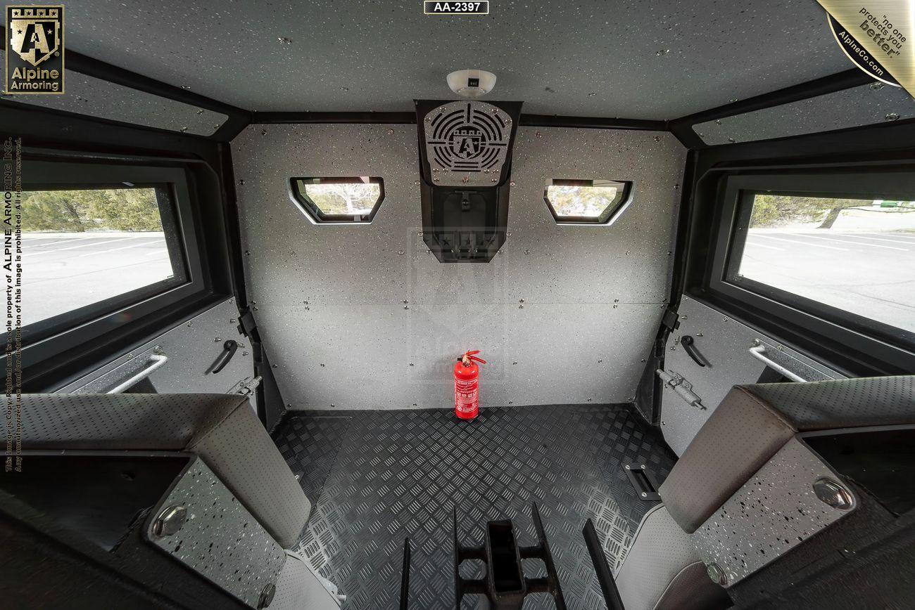 Interior of a bulletproof black Pit-Bull VXT showing gray walls, two small windows, a ventilation system on the ceiling, mounted seats along the sides, and a red fire extinguisher on the floor.