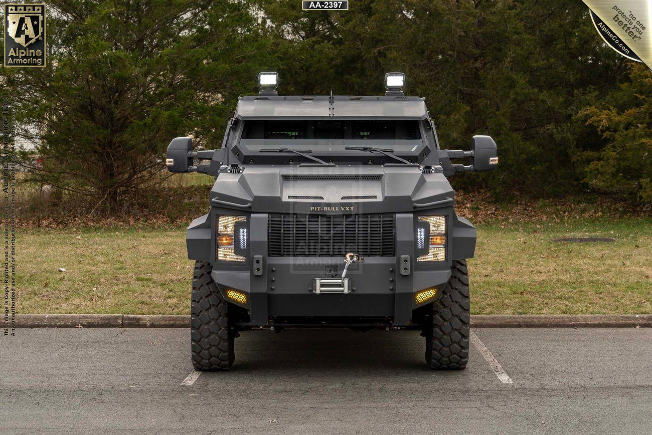 A front view of an Alpine Armoring Pit-Bull VXT armored vehicle parked in an outdoor setting with trees in the background.