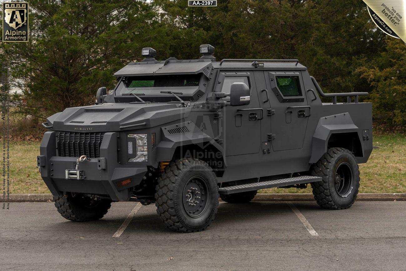 A matte-black armored black Pit-Bull VXT with large tires is parked on asphalt, facing slightly to the left. Trees are visible in the background.