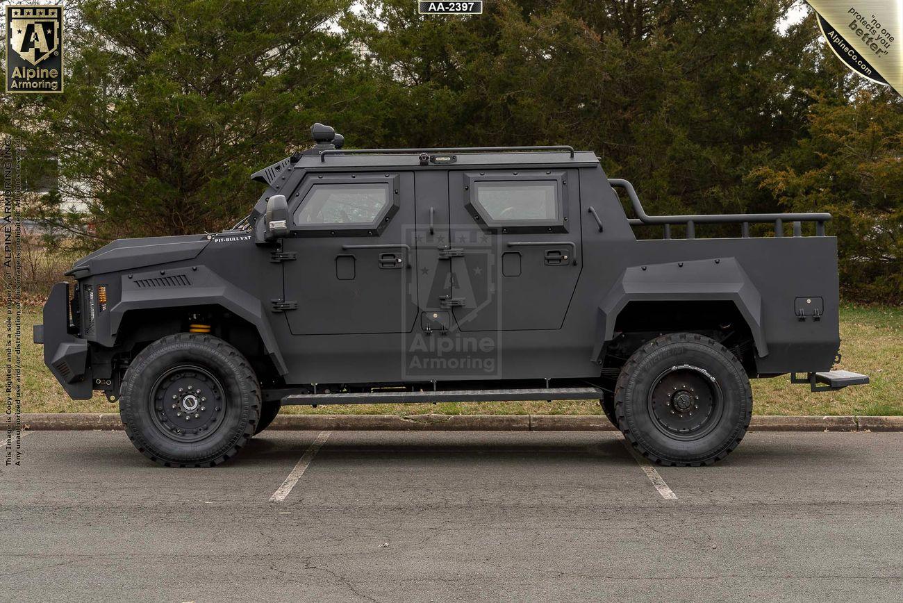 A black armored Pit-Bull VXT is parked on a road with trees in the background. The vehicle has rugged tires and protective plating. The image appears to be from Alpine Armoring.