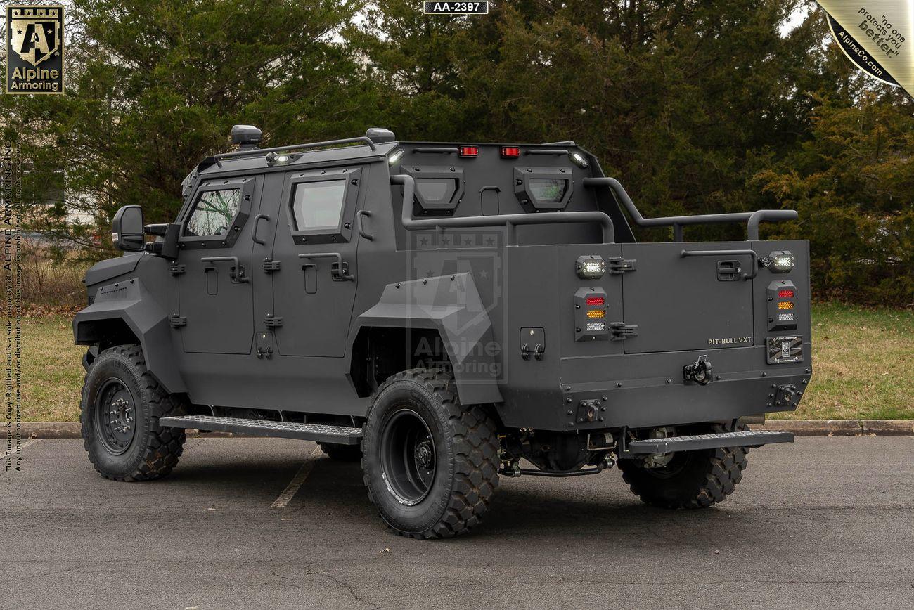 A black armored black Pit-Bull VXT vehicle is parked on a paved road. It has large off-road tires, reinforced windows, and various equipment mounted on its exterior. Trees and grass are visible in the background.