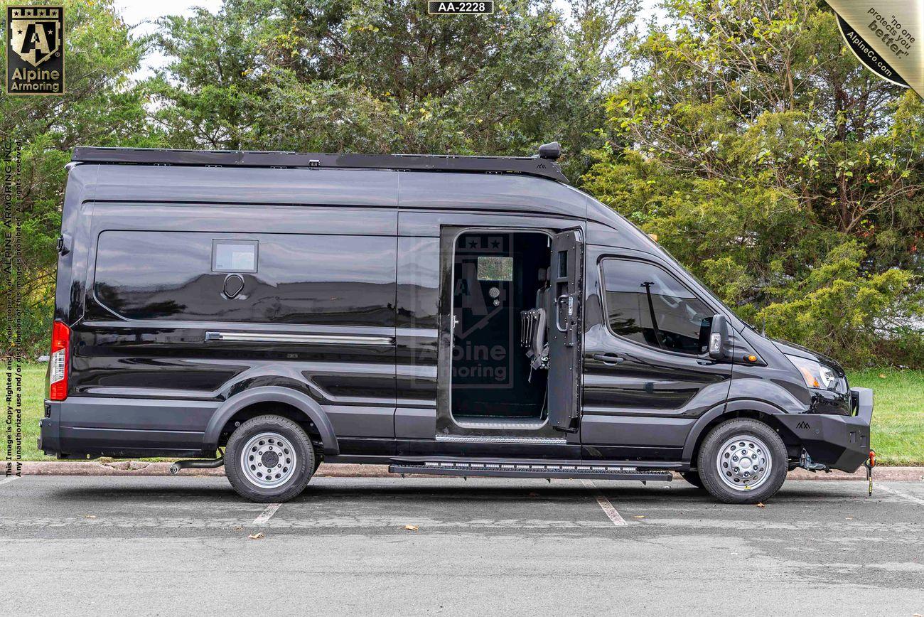 A black Ford Transit Pointer Van is parked on the side of a street with its side door open. The vehicle is surrounded by green trees and a grassy area in the background.