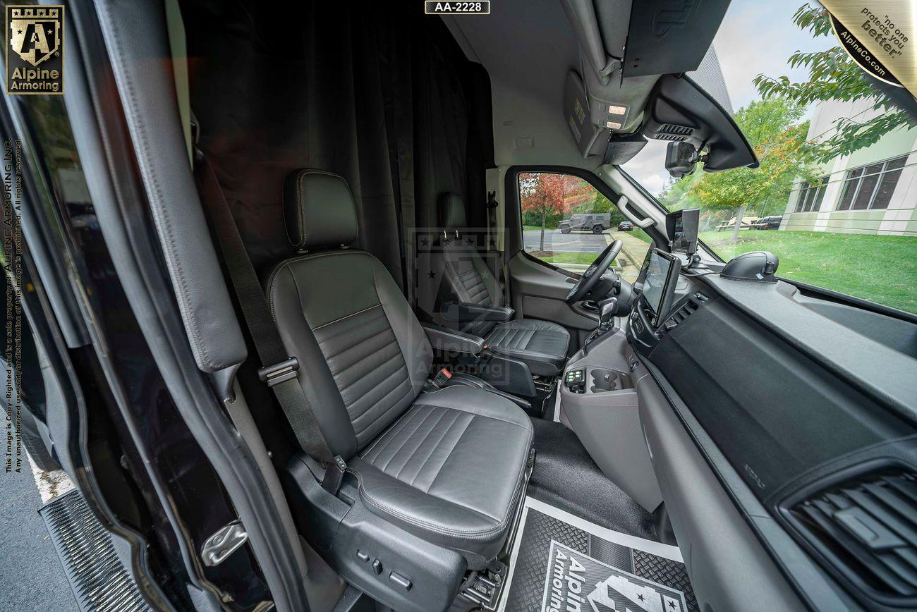 Interior of a Ford Transit Pointer Van showcasing two black leather seats, a central console with a gear shift, and mounted screens on the dashboard. The view includes the windshield with a tree and building outside.