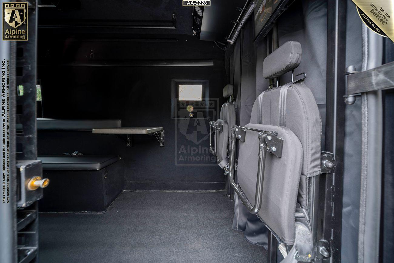 Interior of an armored Ford Transit Pointer Van with seating and a small work desk along the left wall. The seats have metal frames and are attached to the right wall. The environment is enclosed and utilitarian.