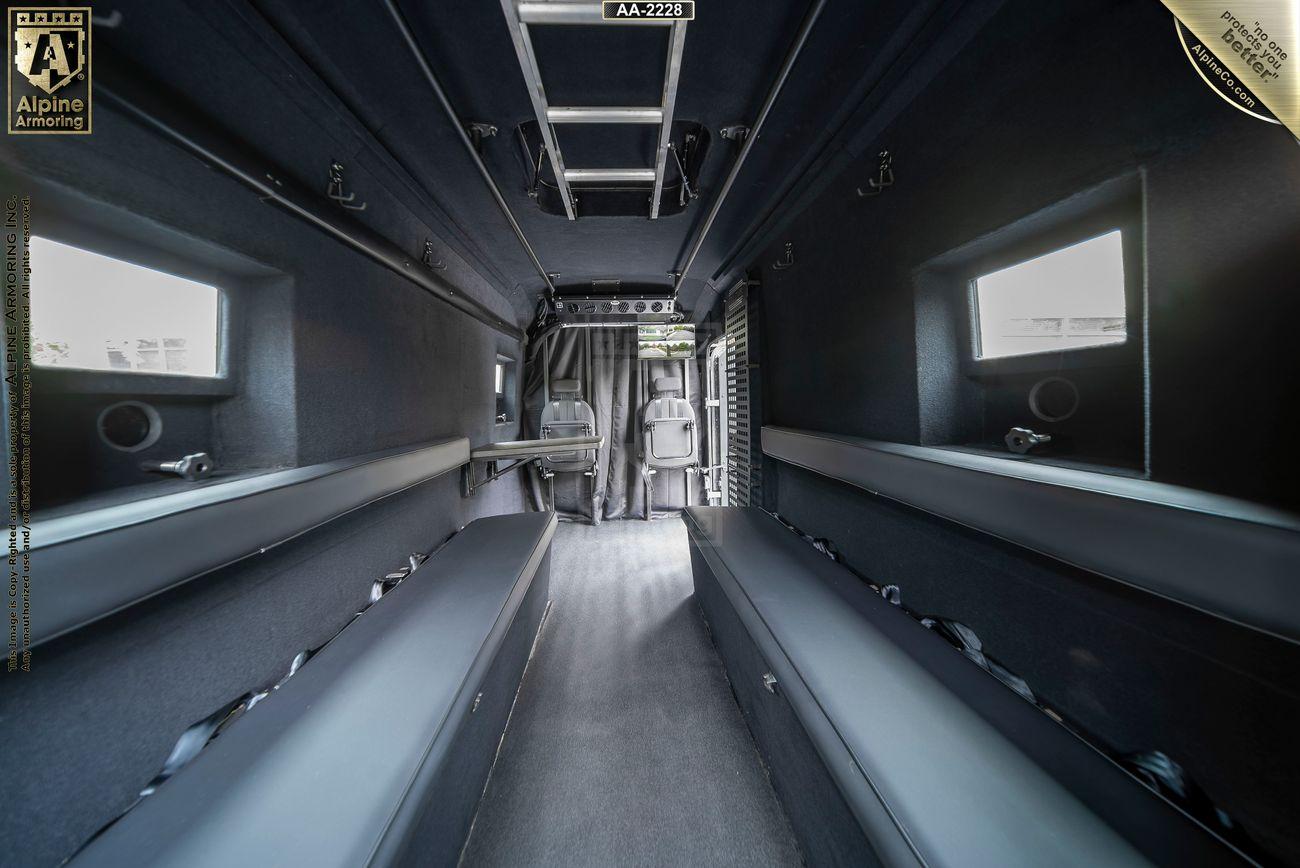 Interior of a dark, armored Pointer Van with long side benches, small windows, and two seats at the front facing a curtained partition.