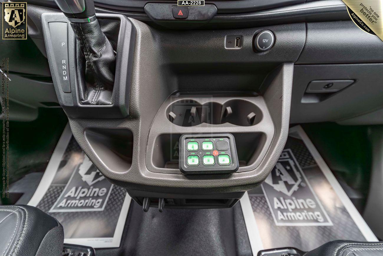 The central dashboard area of a Ford Transit Pointer Van equipped with a control panel, gear shift lever, and cup holders. Branded floormats labeled "Alpine Armoring" are visible beneath the dashboard.