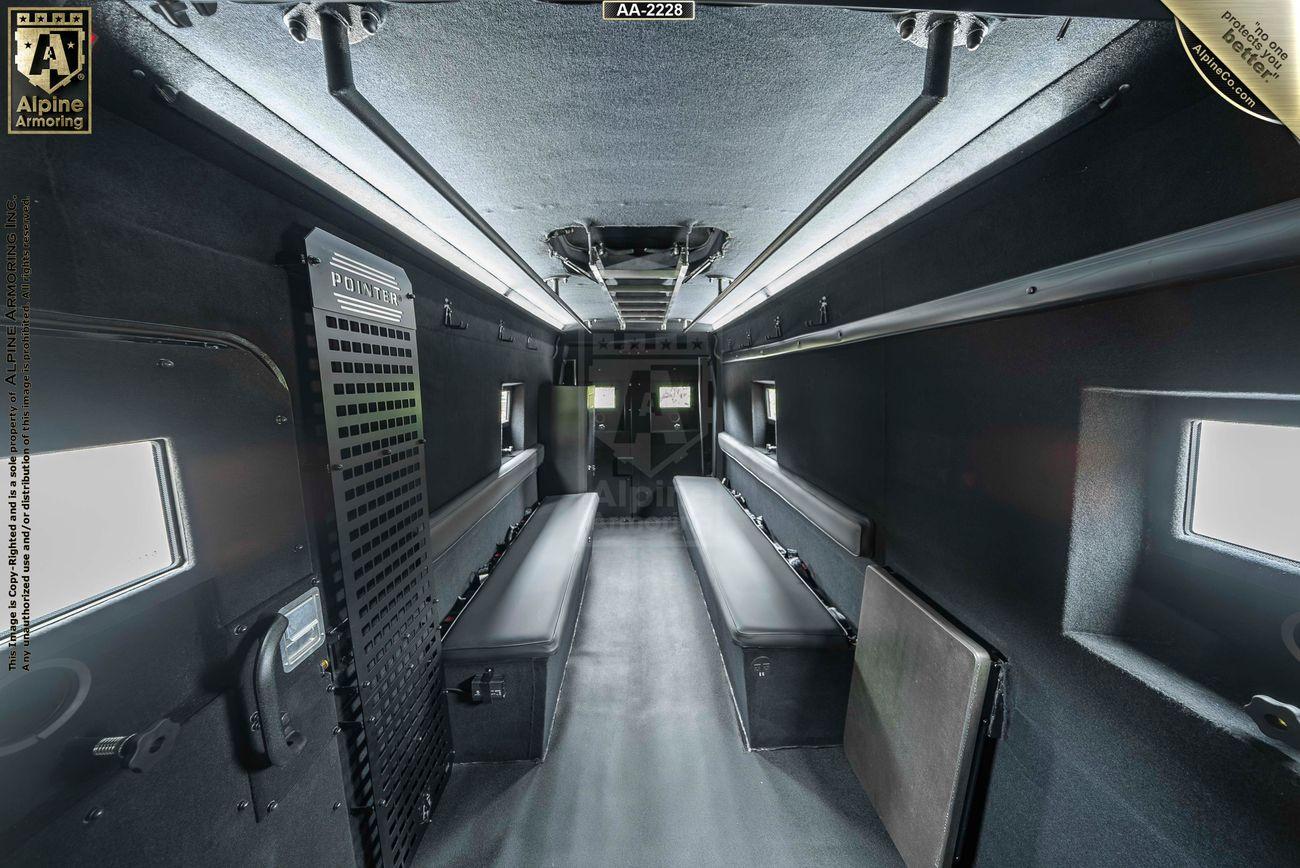 The interior of an armored Pointer Van, featuring metal walls, bench seating along both sides, and small windows, illuminated by overhead lighting.