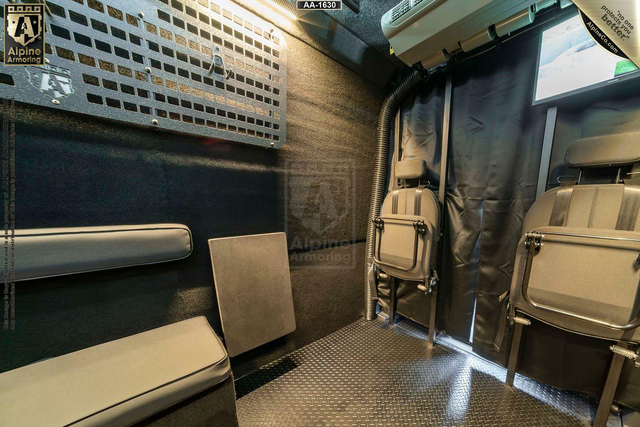 Interior of an armored  Pointer van showing a metal grid wall, a padded bench, and two gray seats with seatbelts. The floor has a diamond plate design.